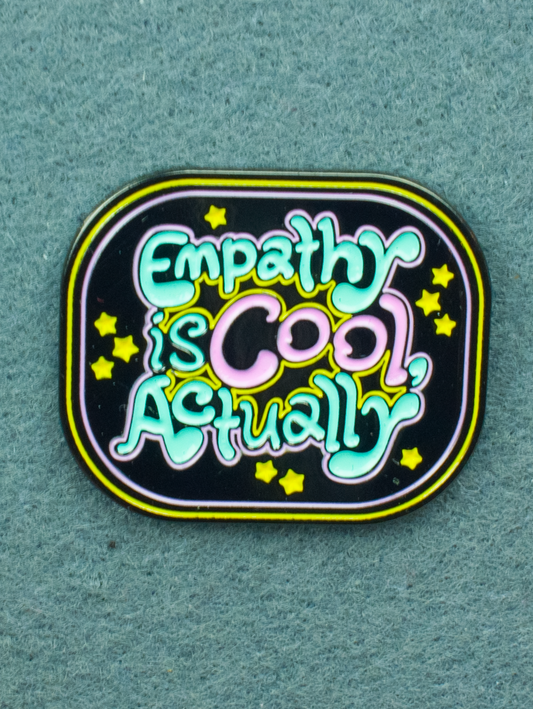 A square enamel pin with rounded corners. Pink and blue text on a black background reads "Empathy is Cool, actually". Several yellow stars are also seen on the design.
