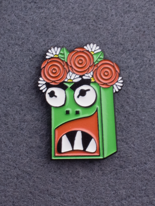 An enamel pin of Garry the kaiju. Green character with white teeth and a flower crown. 
