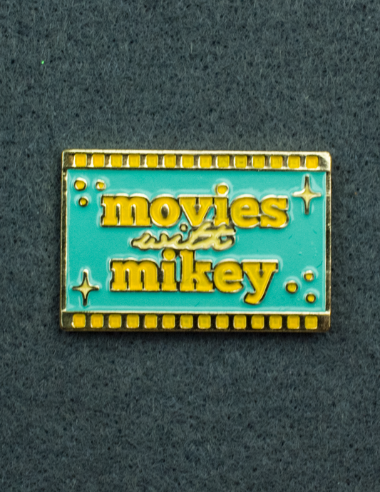 An enamel pin featuring the Movies with Mikey logo on a teal background with yellow film strips on the top and bottom. 